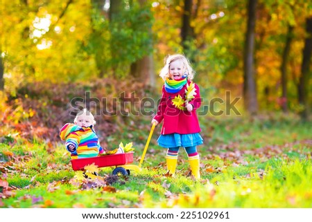 Cute little children, adorable toddler girl and a funny baby boy, brother and sister, playing in a sunny autumn park with a wheel barrow and colorful leaves