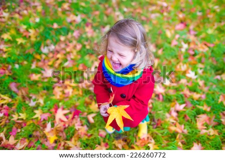 Funny laughing little girl in a red coat and colorful rainbow scarf playing with golden leaves in a beautiful autumn park with maple trees