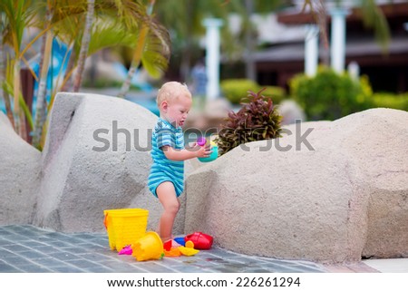 Cute little baby boy playing with plastic toy bucket and watering can at pool side in a tropical resort during summer vacation