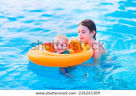 Two brothers, little baby in an orange inflatable ring and his teenager brother, playing together in a pool learning to swim