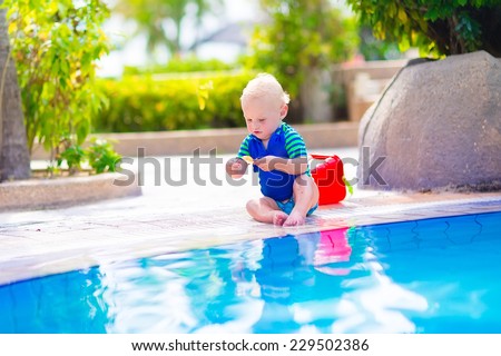 Cute funny baby boy playing with red toy bucket sitting at a swimming pool in a tropical resort on a hot summer day