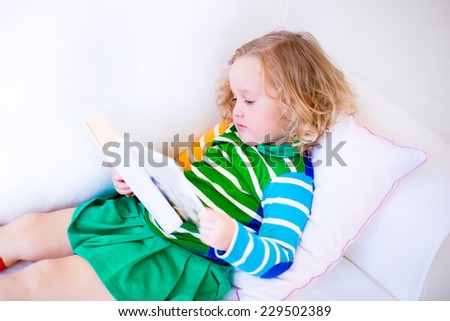 Happy laughing little girl reading a book relaxing on a white couch at home