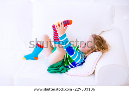 Funny little girl trying to put on warm socks on a white couch at home