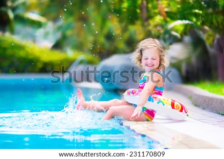 Adorable little girl with curly hair wearing a colorful swimming suit playing with water splashes at beautiful pool in a tropical resort having fun during family summer vacation