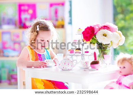 Adorable toddler girl with curly hair wearing a colorful dress on her birthday playing tea party with a doll, toy dishes, cup cakes and muffins in a sunny room with window