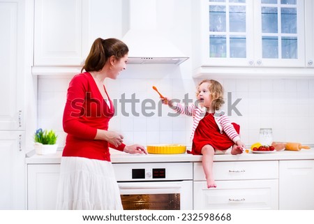 Young mother and her adorable daughter, cute funny toddler girl in a red dress, baking a pie together preparing healthy lunch in a white sunny kitchen