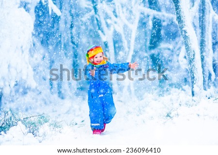 Happy laughing child, cute toddler girl in a colorful snowsuit and hat, running in a snowy winter park catching snow flakes