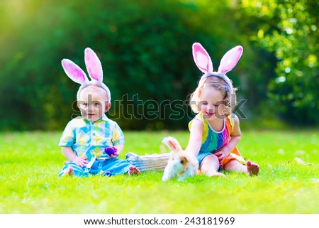 Two little children, cute curly toddler girl and funny baby boy wearing bunny ears having fun at Easter egg hunt playing with basket and toy rabbit in a sunny spring garden