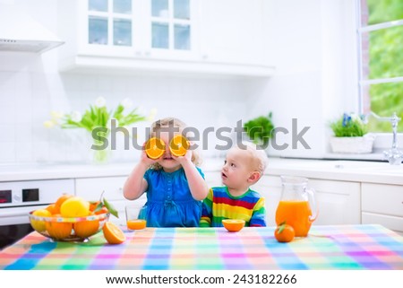 Cute funny little girl and adorable baby boy drinking freshly squeezed orange juice for healthy breakfast in a white kitchen with window on a sunny summer morning