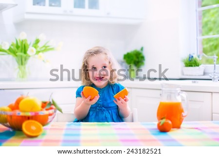 Cute funny little girl drinking freshly squeezed orange juice for healthy breakfast in a white kitchen with window on a sunny summer morning