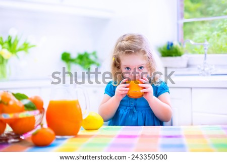 Cute funny little girl drinking freshly squeezed orange juice for healthy breakfast in a white kitchen with window on a sunny summer morning