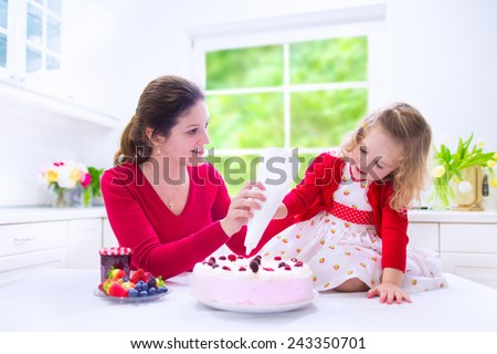 Happy family, young mother and her adorable little daughter, cute curly toddler girl in a red dress, baking fresh strawberry cream cake with fruit and berry in a white kitchen on a sunny summer day