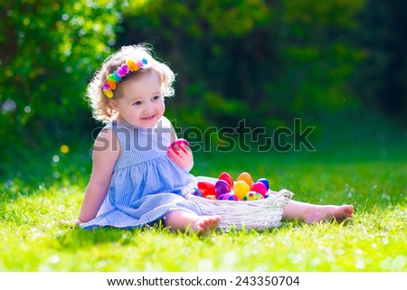 Cute little toddler girl with curly hair wearing a blue summer dress having fun during Easter egg hunt relaxing in the garden on a sunny spring day