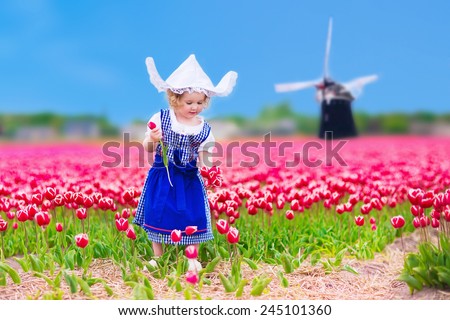 Adorable curly toddler girl wearing Dutch traditional national costume dress and hat playing in a field of blooming tulips next to a windmill in Amsterdam region, Holland, Netherlands