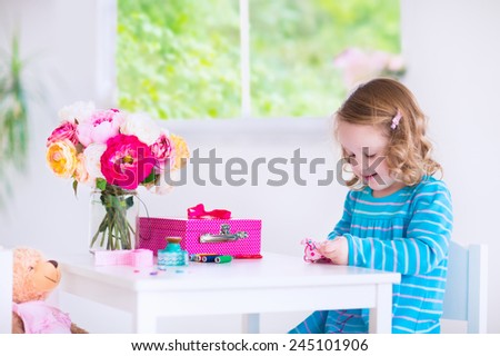 Cute creative little girl sewing a dress for her teddy bear doll, playing with needles and ribbons in a white sunny room at home or preschool