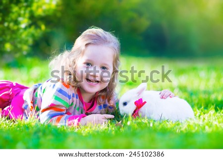 Adorable curly toddler girl playing with a real rabbit in a sunny summer garden, child feeding bunny a carrot.