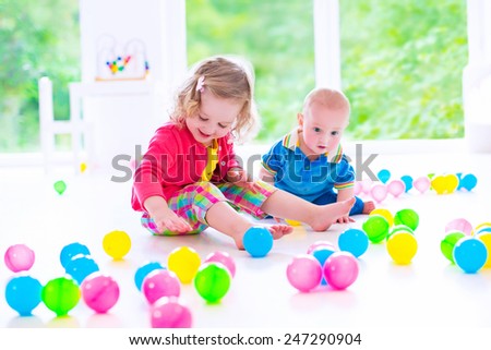 Two happy little children, cute curly toddler girl and a funny baby boy playing together with colorful balls in a white sunny room with big window
