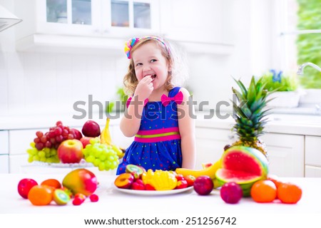Cute curly little girl in a colorful summer dress eating fresh tropical fruit and berry for healthy breakfast snack in a white sunny family kitchen
