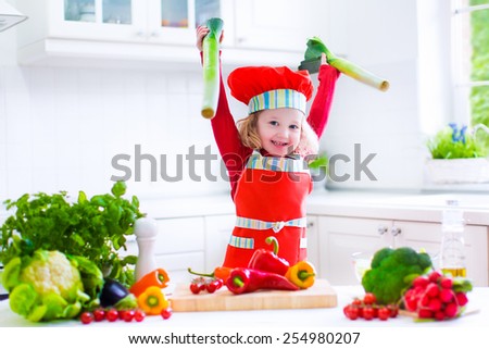 Happy little girl in a red chef hat and apron preparing healthy lunch with fresh raw vegetables, making salad with tomato, eggplant, pepper and cauliflower in a white kitchen at home