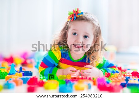 Cute funny preschooler little girl in a colorful shirt playing with construction toy blocks building a tower in a sunny kindergarten room
