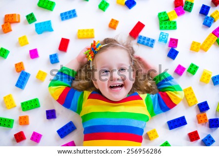 Cute funny preschooler little girl in a colorful shirt playing with construction toy blocks building a tower in a sunny kindergarten room