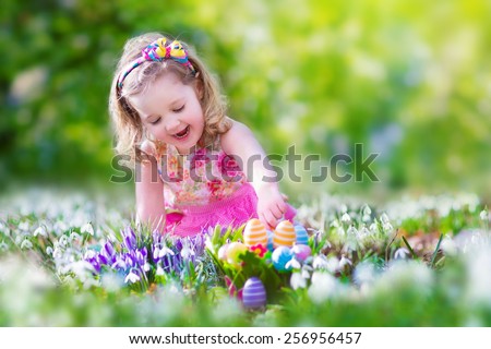 Adorable curly toddler girl in a pink summer dress playing with Easter eggs during egg hunt in a sunny garden with first white spring flowers