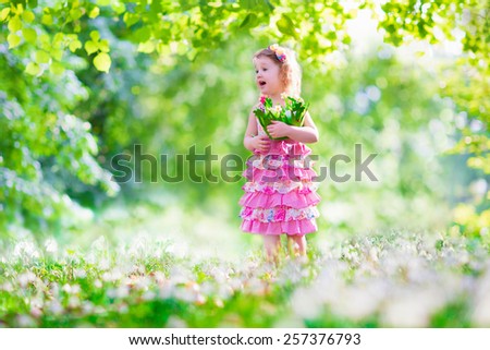 Adorable curly toddler girl in a pink summer dress playing with Easter eggs during egg hunt in a sunny garden with first white spring flowers
