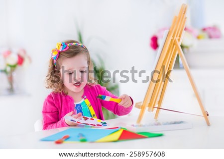 Cute happy little girl, adorable preschooler, painting with water color on canvas standing on a wooden easel in a sunny white room at home or elementary school, creative young artist at work