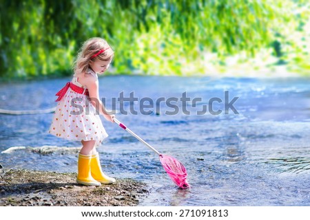 Child playing in a river. Cute little girl in summer dress and rain boots catching fish and frog with a colorful net standing in water. Kids play outdoors. Young explorer and fisherman in wild nature.