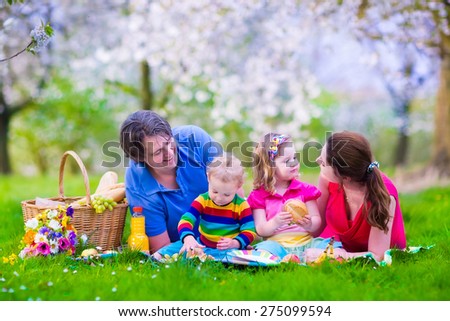 Young family with kids having picnic outdoors. Parents with two children relax in blooming summer garden. Mother, father, little girl and baby boy eat sandwich and fruit, drink juice for healthy lunch