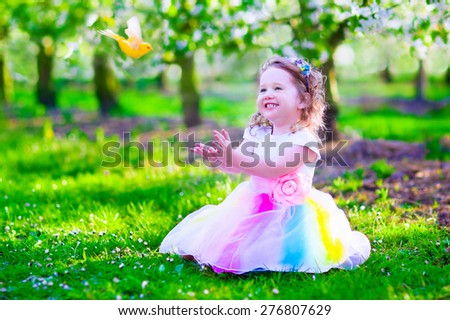 Child playing with a bird. Happy laughing little girl in fairy costume with wings feeding a pet parrot in a cherry tree garden holding a bird cage. Kids having fun in blooming fruit orchard in spring.