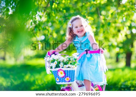 Happy child riding a bike. Cute kid biking outdoors. Little girl in a blue dress on a pink bicycle with daisy flowers in a basket. Healthy preschool children summer activity. Kids playing outside.