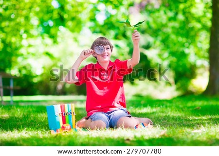 Happy school child, laughing boy sitting on a green lawn in the school yard looking through a magnifying glass exploring leaves and reading books for science class, back to school concept
