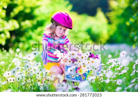Happy child riding a bike. Cute kid in safety helmet biking outdoors. Little girl on a pink bicycle with daisy flowers in a basket. Healthy preschool children summer activity. Kids playing outside.
