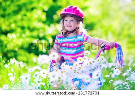 Happy child riding a bike. Cute kid in safety helmet biking outdoors. Little girl on a pink bicycle with daisy flowers in a basket. Healthy preschool children summer activity. Kids playing outside.