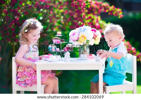 Tea garden party for kids. Child birthday celebration. Little boy and girl play outdoor drinking hot chocolate and eating cake. Children eat sweets. Kid event with toy dish and flower decoration.