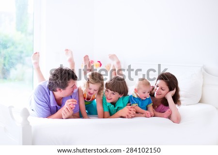 Young family with three kids at home in bed. Parents with children relaxing in bed on a sunny morning. Mother, father, baby boy, toddler girl and school child together. White interior and bedding.
