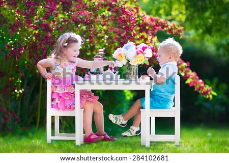 Tea garden party for kids. Child birthday celebration. Little boy and girl play outdoor drinking hot chocolate and eating cake. Children eat sweets. Kid event with toy dish and flower decoration.