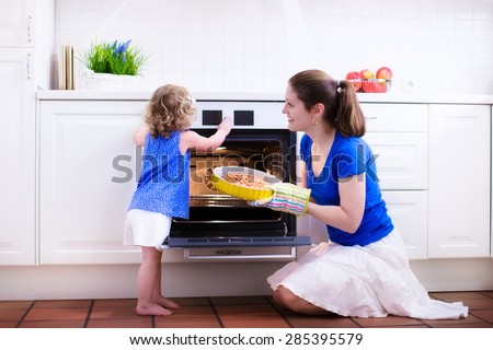 Mother and child bake a pie. Young woman and her daughter cook in a white kitchen. Kids baking pastry. Children helping to make dinner. Modern interior with oven and other appliances. Family eating.