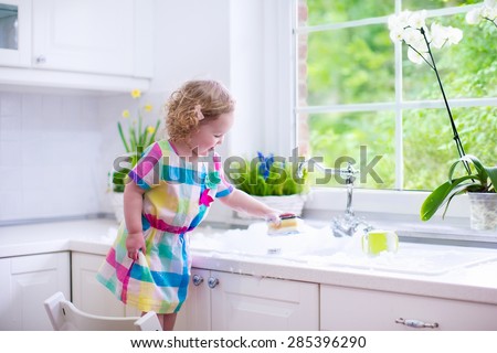 Child washing dishes. Kids wash plates and cups. Little girl helping in the kitchen playing with water and foam in a white sink with retro tap. Chores for children. Modern home interior with window.