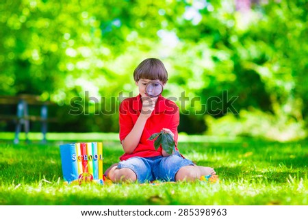 Happy school child, laughing boy sitting on a green lawn in the school yard looking through a magnifying glass exploring leaves and reading books for science class, back to school concept
