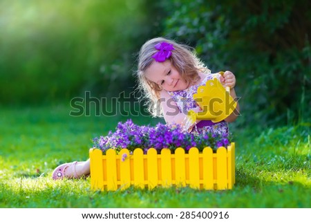 Child working in the garden. Kids gardening. Children watering flowers. Little girl with water can on a green lawn in the backyard in summer. Toddler kid playing outdoors planting purple flower pots.