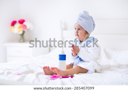 Child after bath. Cute little girl with wet curly hair wearing a bathrobe and head towel sitting on a white bed using lotion and brush. Hygiene for kids. Bathroom textile for babies and children.