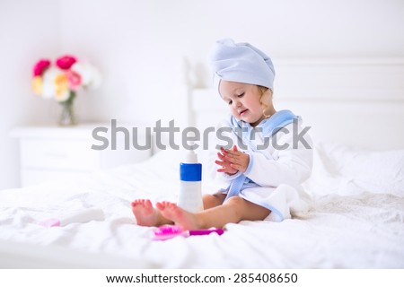 Child after bath. Cute little girl with wet curly hair wearing a bathrobe and head towel sitting on a white bed using lotion and brush. Hygiene for kids. Bathroom textile for babies and children.