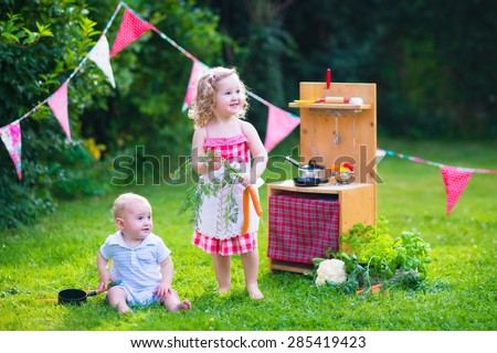 Kids playing with a toy kitchen. Children play in a summer garden. Boy and girl cooking and baking healthy vegetables in the backyard. Toddler child and baby cook together. Outdoor fun in summer.