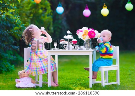 Garden birthday party for children. Kids outdoor celebration. Little boy and girl drinking tea and eating cake playing in the backyard in summer. Toddler and baby play with toy dishes and eat cupcakes