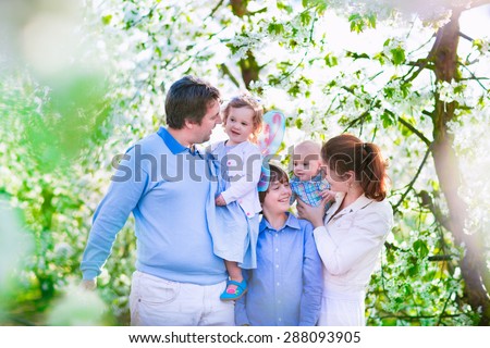Family with kids in a blooming spring garden. Parents with three children, baby, toddler girl and boy enjoying picnic on a farm with apple and cherry trees. Mother, father, son and daughter in summer
