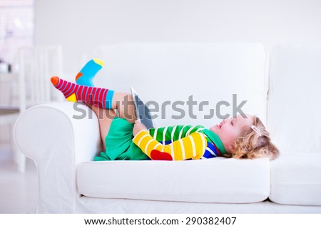 Little girl holding tablet pc relaxing on a white couch. Kids using computer at home or preschool. Children learning with digital devices. Child playing online game. Toddler kid and modern gadget.