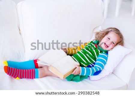 Little girl reading a book relaxing on a white couch. Kids read books at home or preschool. Children learning and doing homework after school. Child playing. Toddler kid in colorful dress on a sofa.
