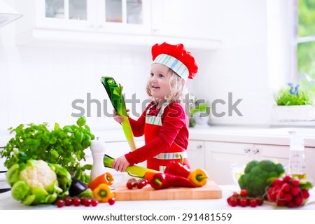 Kids cooking fresh vegetable salad in white kitchen. Children cook vegetables for vegetarian lunch. Toddler in chef hat eats healthy dinner. Little girl preparing and eating raw meal. Child nutrition.